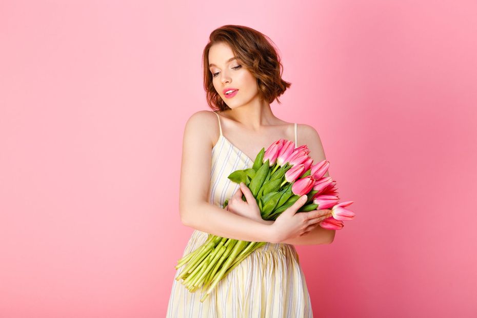 Una persona con cabello ondulado hasta los hombros, vestida con un vestido blanco de verano, sostiene un ramo de tulipanes rosas sobre un fondo rosa.
