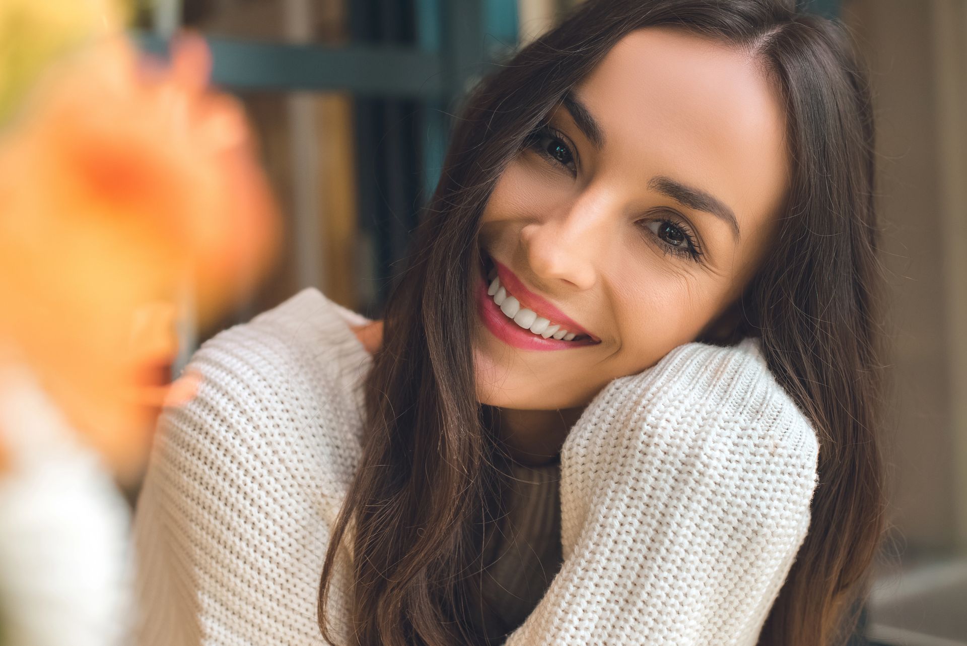 Una mujer sonriente con un suéter de punto blanco, apoyando la barbilla en la mano, con un objeto naranja borroso en primer plano.