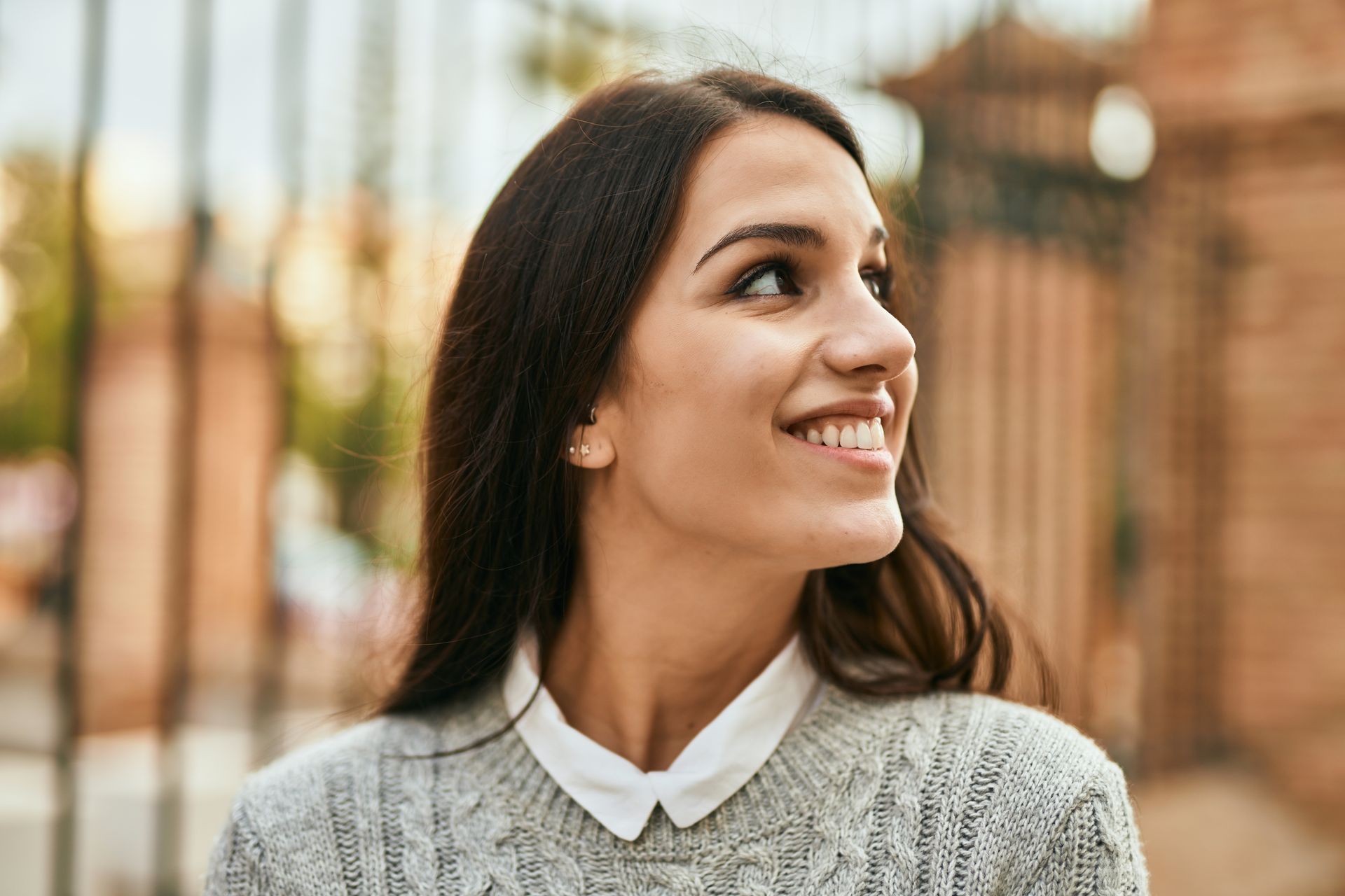 Una persona sonriente con cabello largo y oscuro lleva un suéter gris sobre una camisa blanca con cuello mientras mira fuera de cámara al aire libre.