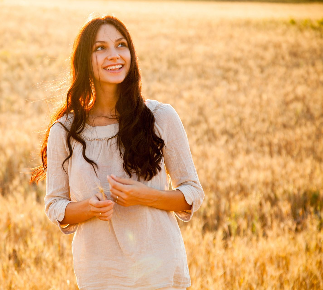 Una persona con cabello largo y oscuro sonríe mientras está de pie en un campo de trigo dorado al atardecer.