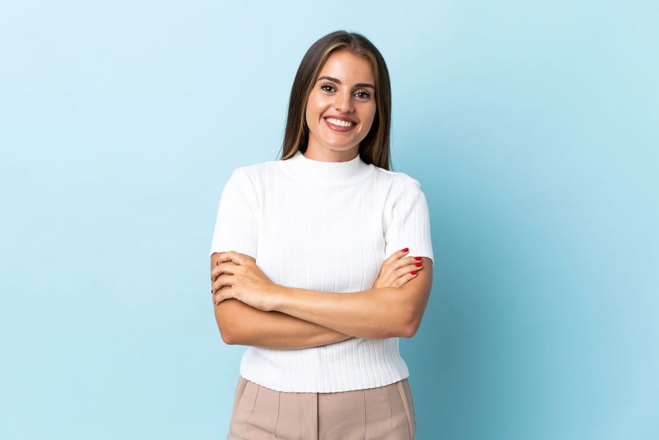 Una persona sonriente, con cabello castaño largo y vestida con una camisa blanca texturizada, permanece de pie con los brazos cruzados sobre un fondo azul.