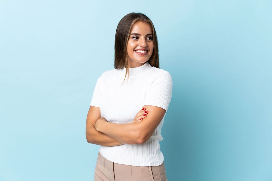 Una persona con cabello castaño largo, vestida con una blusa blanca y pantalones color canela, sonríe con los brazos cruzados sobre un fondo azul claro.