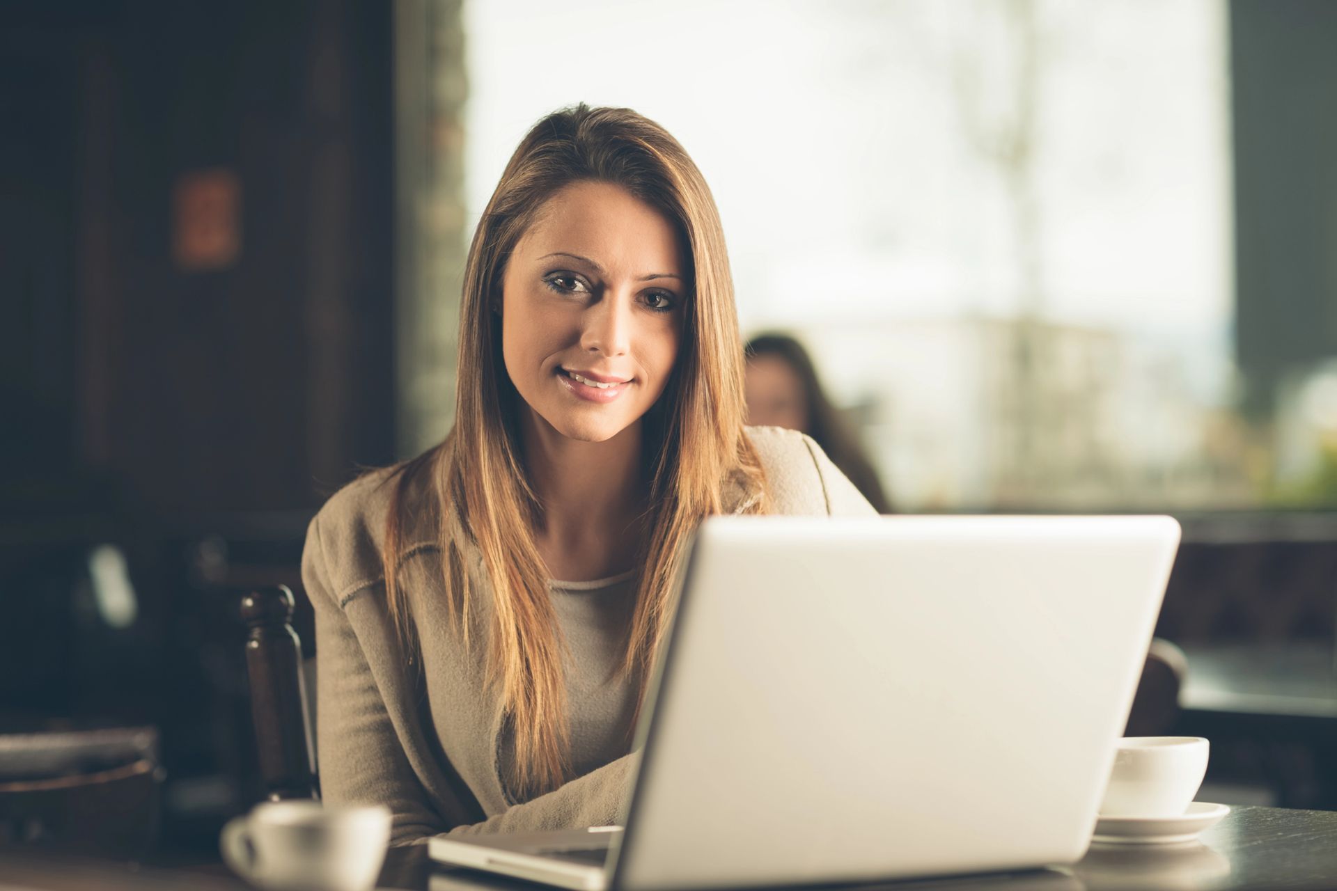 Una persona con cabello largo sentada en una mesa con una computadora portátil, sonriendo a la cámara en un ambiente de cafetería.