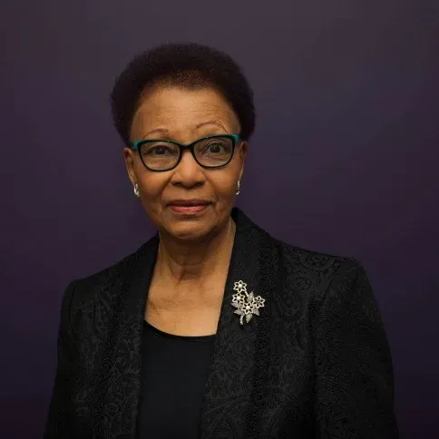 A woman wearing glasses and a black jacket is standing in front of a purple wall.