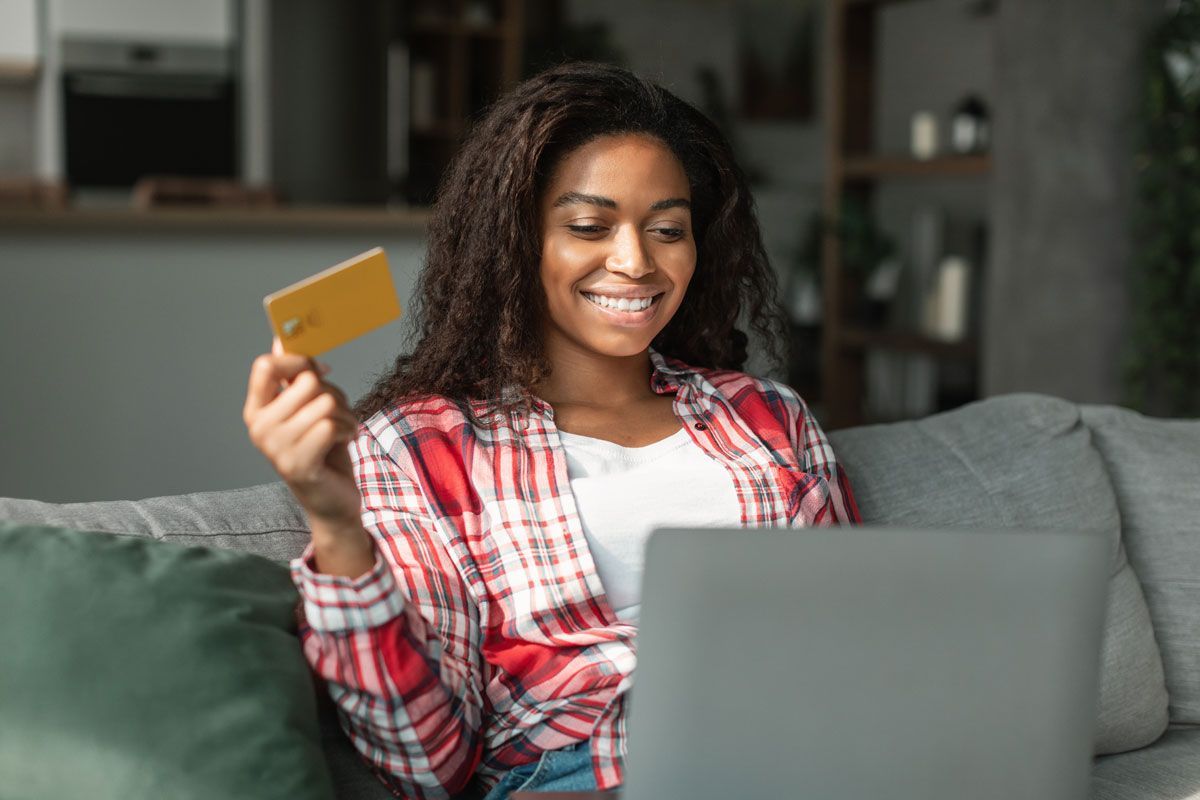 Woman holding a gold credit card, smiling, using a laptop, sitting on a couch.