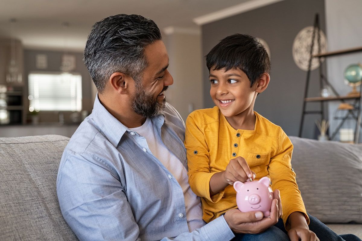 Man and boy putting a coin in a piggy bank while seated on a sofa.