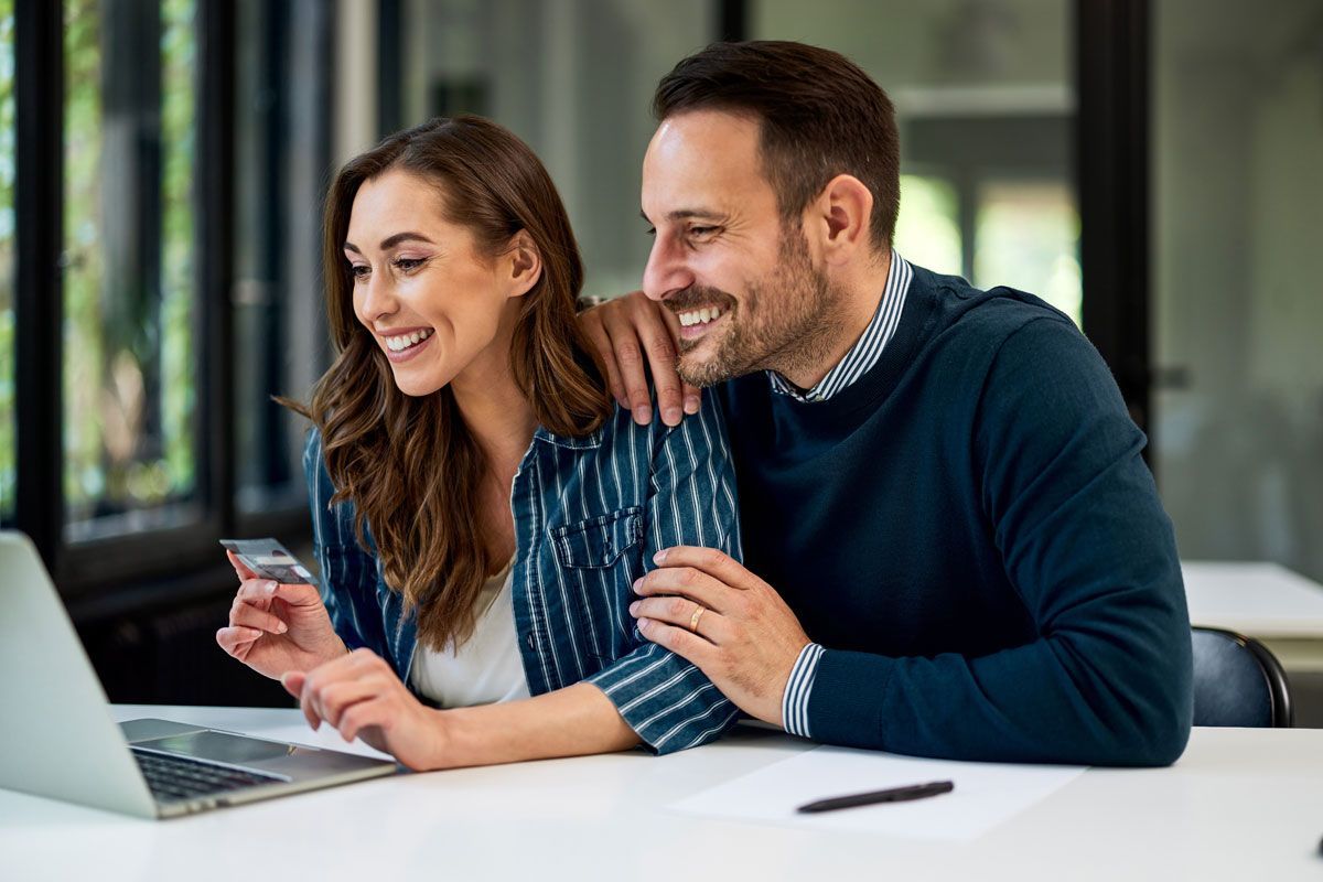 Couple happily shopping online together, smiling at a laptop screen. Woman holds a credit card.