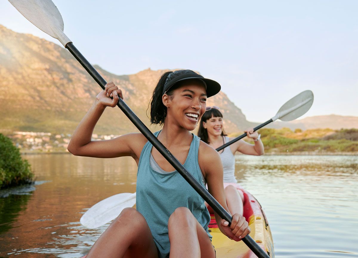 Two people kayaking on a calm lake, smiling. Mountains in the background, sunny.