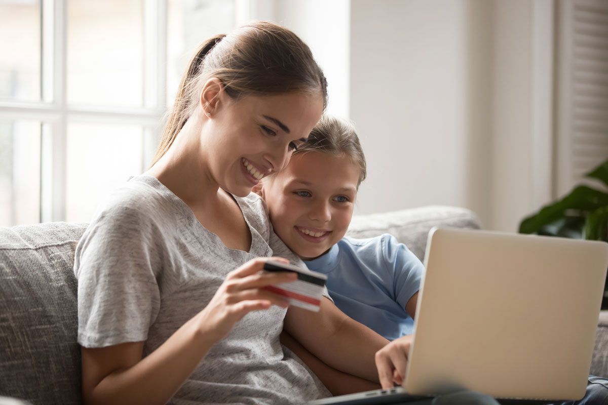 Woman and child smile while looking at a laptop and holding a credit card on a couch.