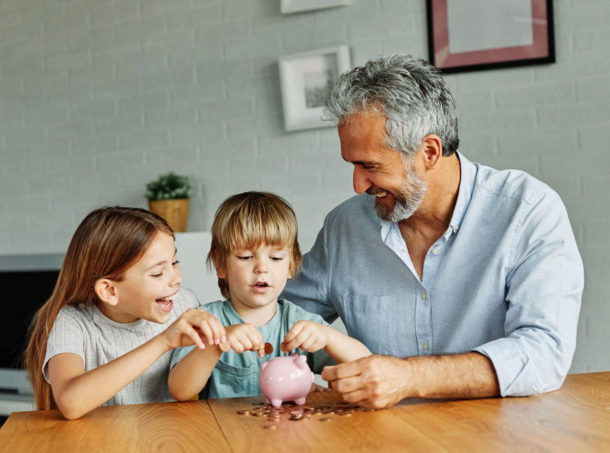 Man and two children putting coins into a pink piggy bank on a wooden table.