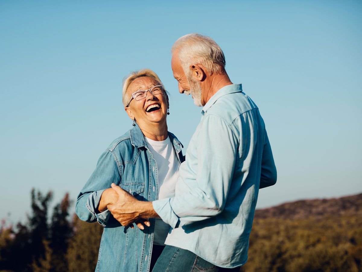 Smiling elderly couple dancing outdoors in front of a blue sky and trees.