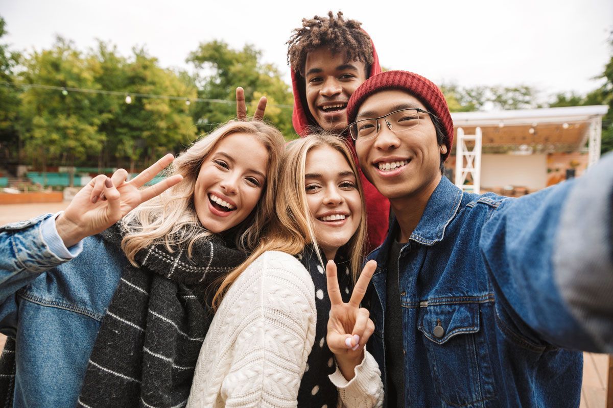 Five smiling friends take a selfie outdoors; peace signs, red beanie, denim jackets.