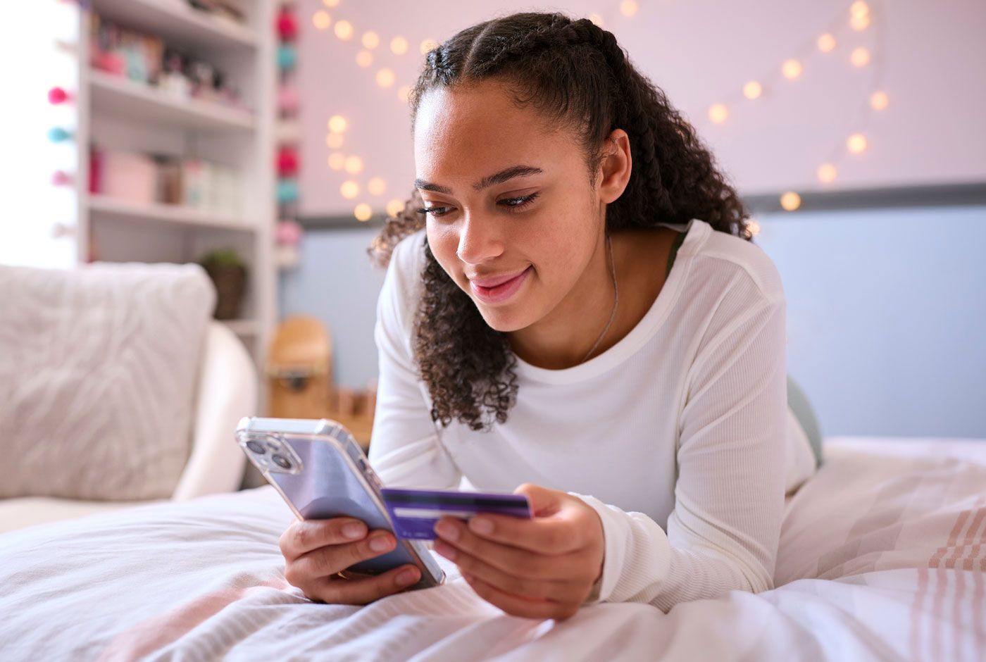 Young person looking at a phone while holding a credit card on a bed in a bedroom.
