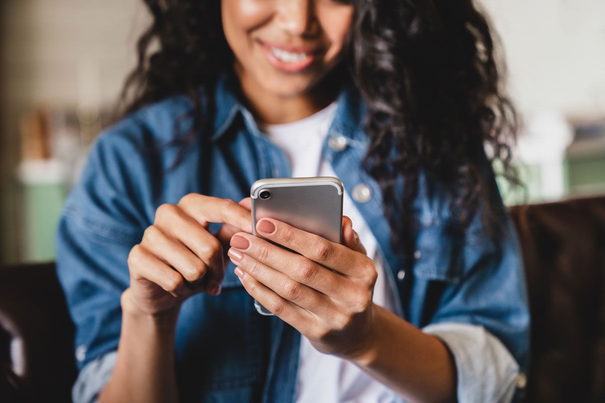 Woman smiling, using a smartphone, wearing a blue denim shirt.
