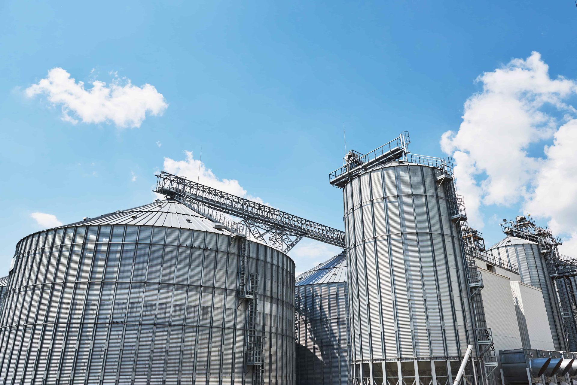 Tall, metallic grain silos under a bright blue, cloudy sky, connected by an industrial metal conveyor structure.