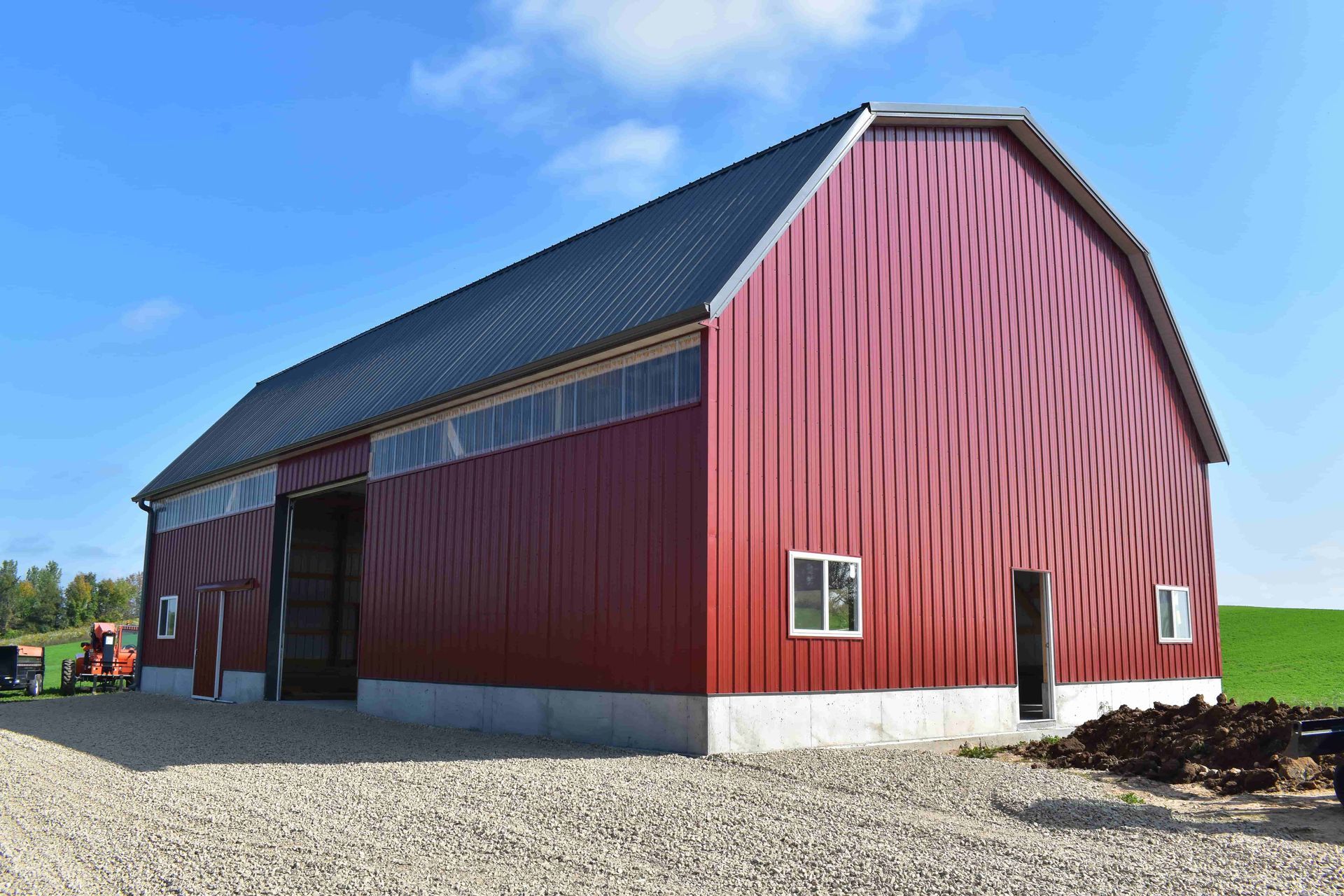 A large, red, gambrel-roof barn with a concrete foundation stands in a rural landscape under a clear blue sky.