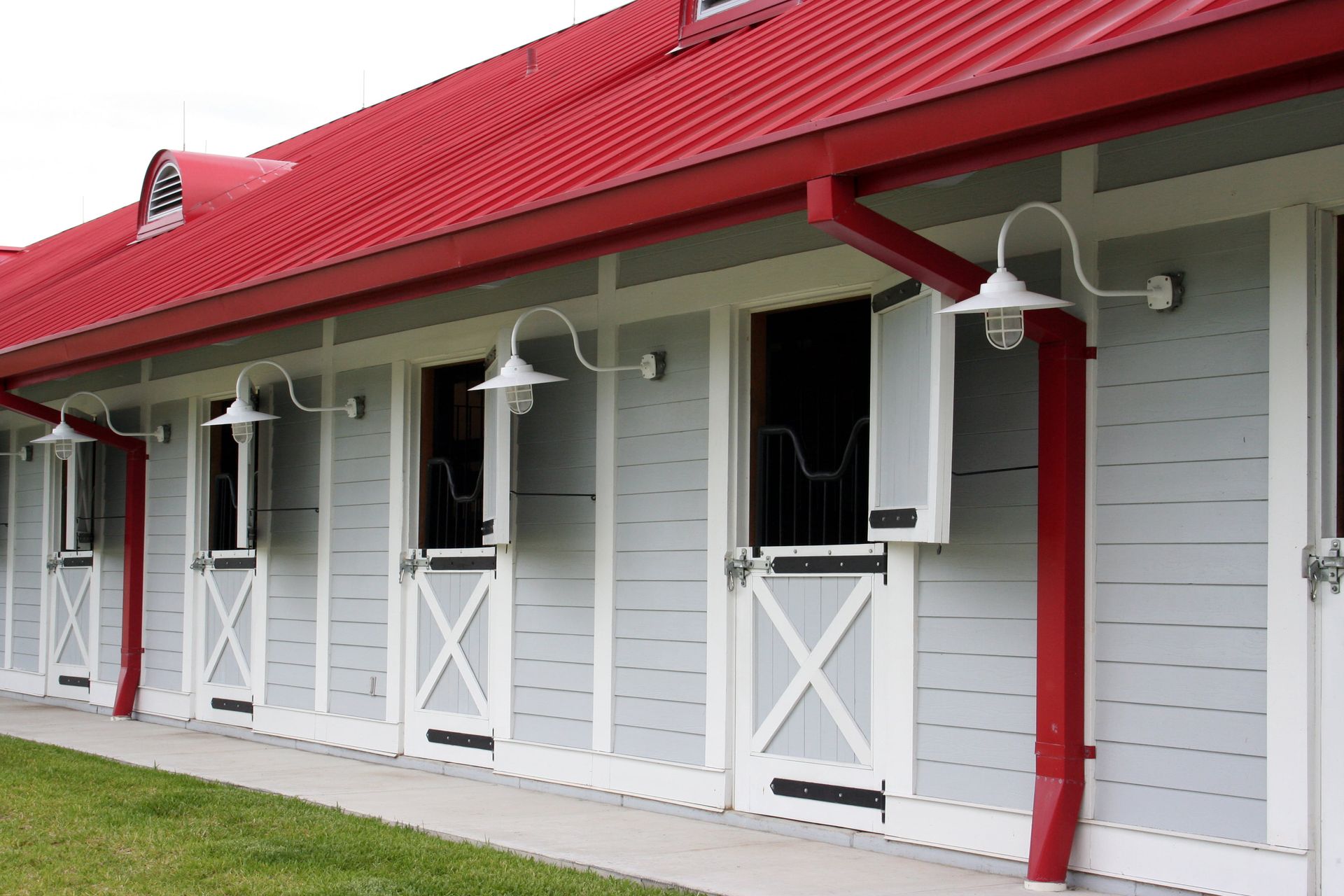 A row of horse stalls featuring gray wood-paneled walls, white trim, red roofs, and decorative gooseneck outdoor lighting.