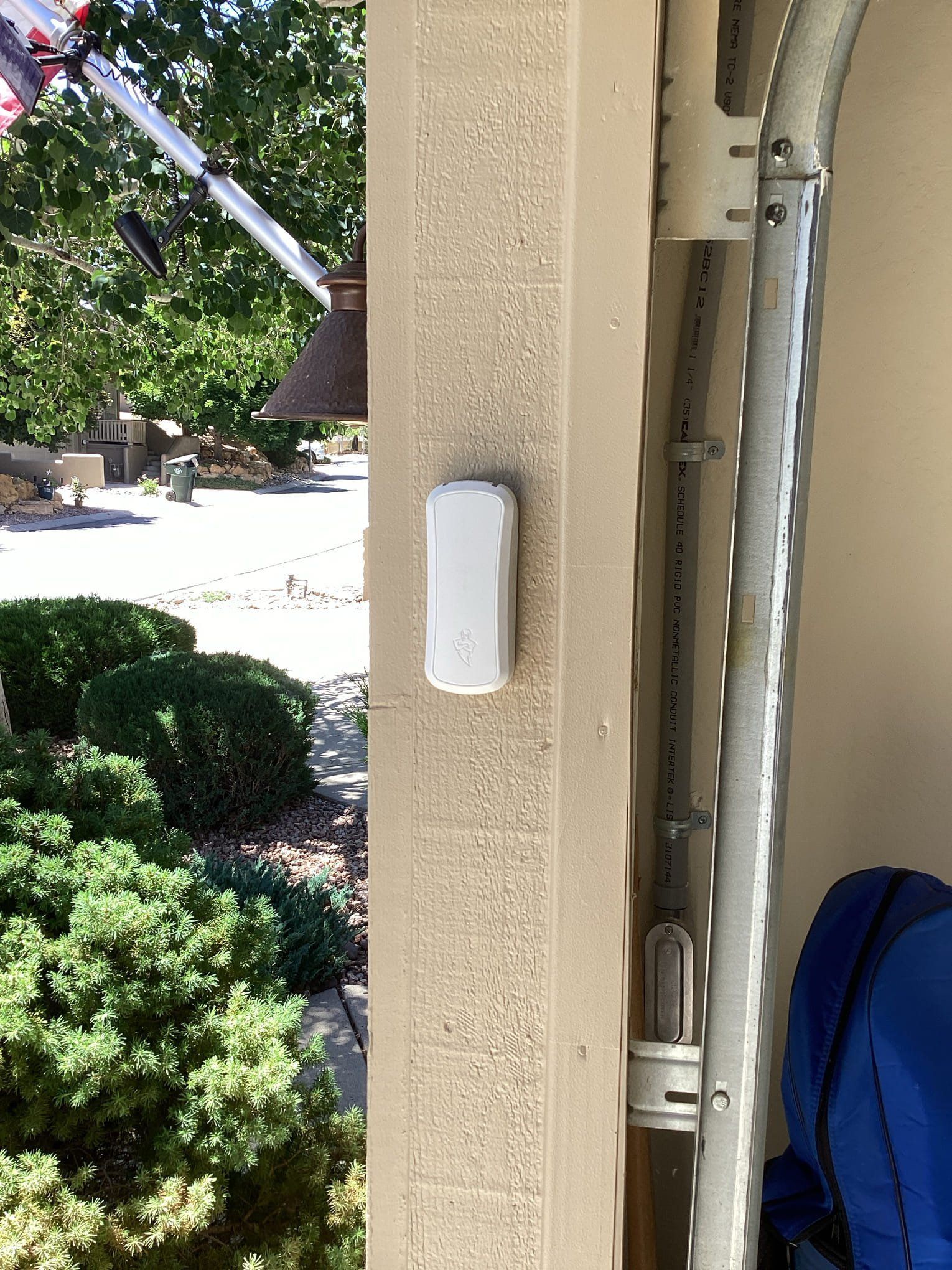 White sensor on a tan pillar beside a garage door. Outdoors, a street and greenery are visible.