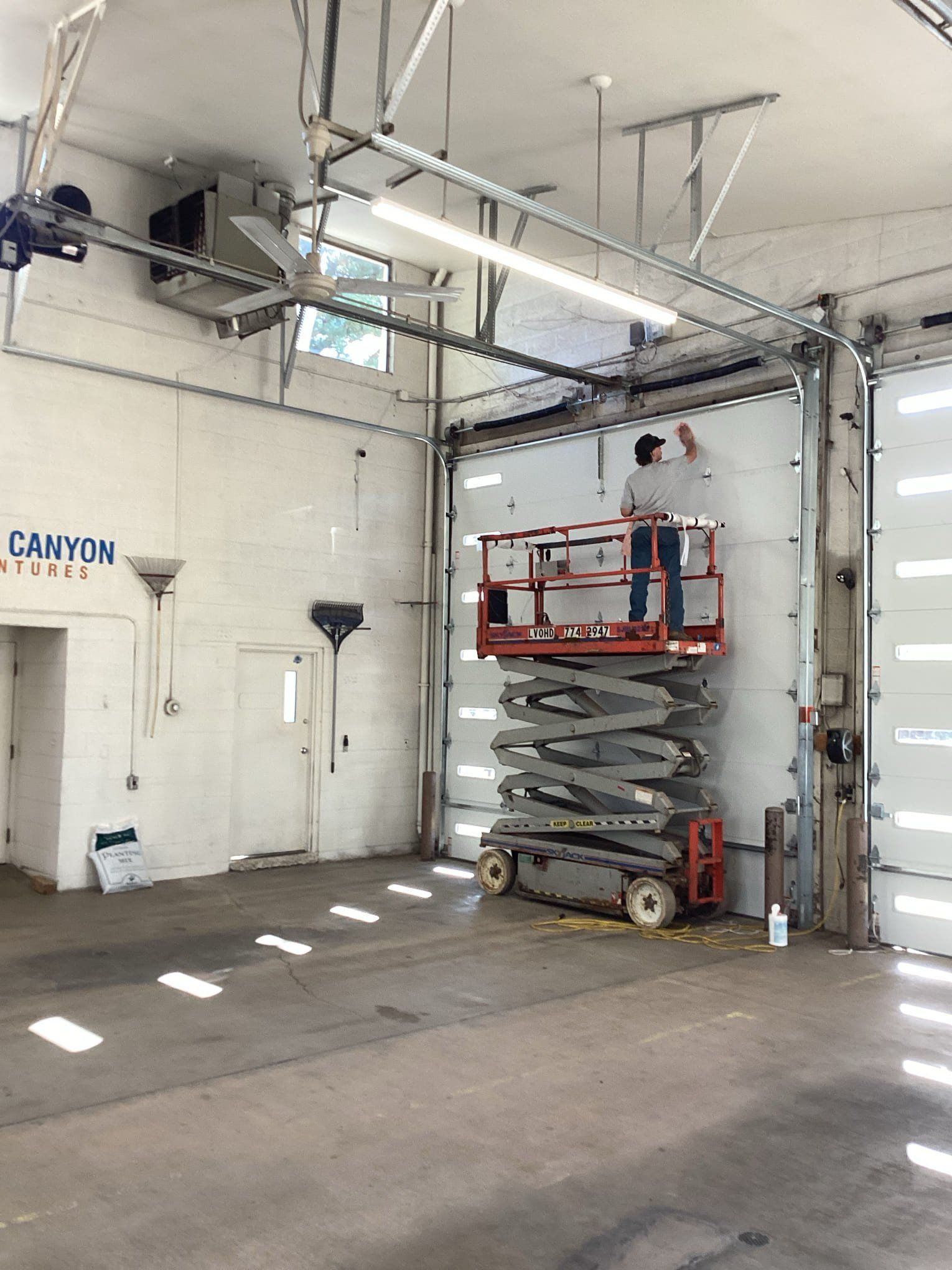 A person on a scissor lift is working on a garage door inside a garage. The garage has white walls and a metal door.