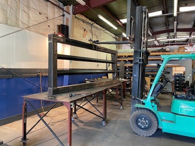 A large metal frame being lifted by a teal forklift inside a workshop. The frame sits on a red metal table with wheels.