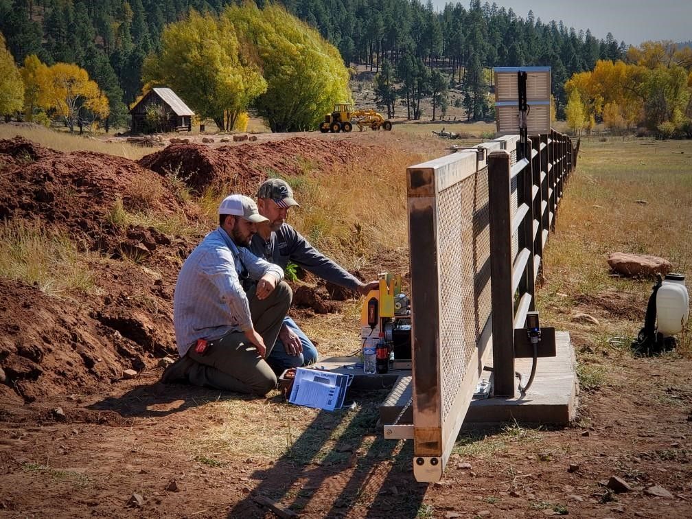 Two men crouch near a scientific instrument in a rural field, analyzing data near a wooden fence and a small building.