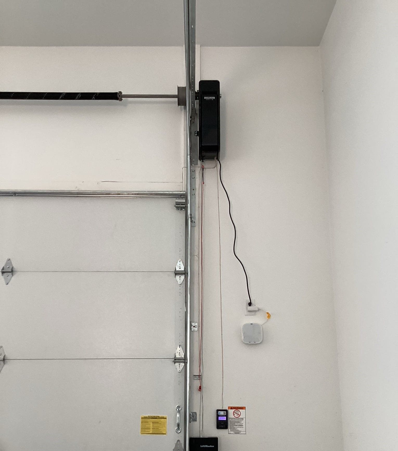 Garage door opener installed on a white wall, with a black motor housing and a wall-mounted control panel.