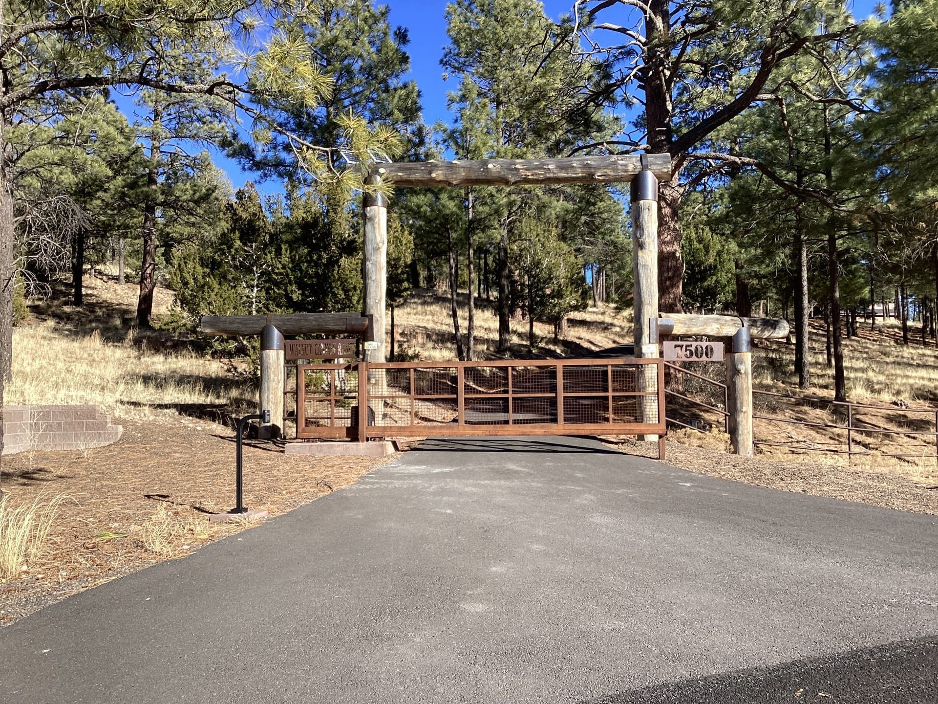 A gated entrance to a property with a brown metal gate and a wooden archway. The setting is a wooded area with a paved driveway.
