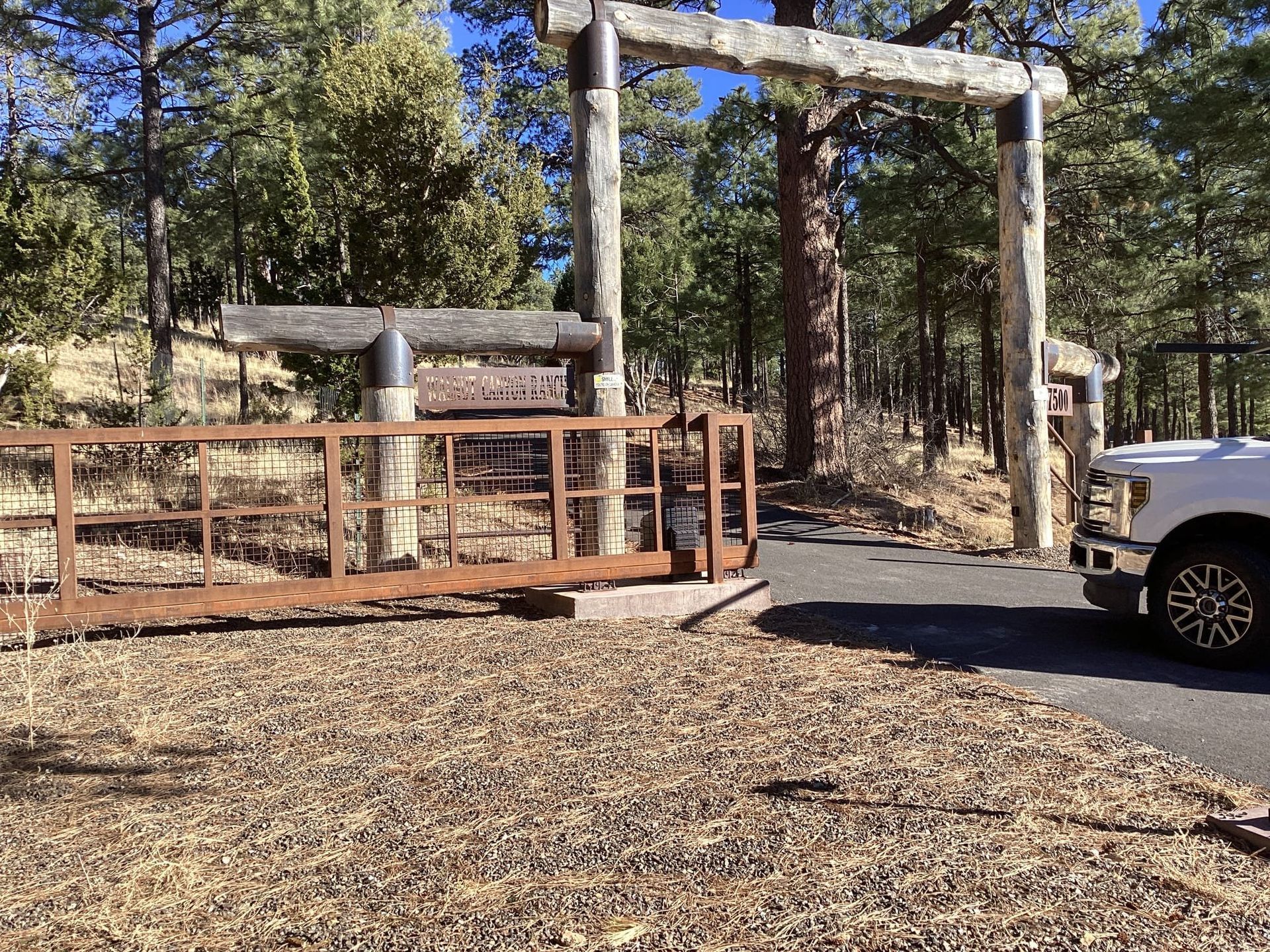 Gate and archway entrance to a property; a brown rusty gate, a white truck, and trees in the background.