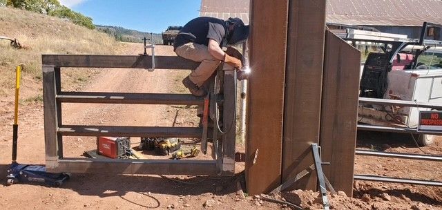 A person welds a metal gate to a post outdoors on a sunny day. They are perched on top of the gate.
