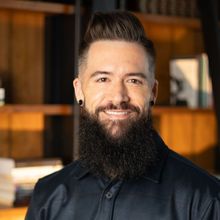 Man with dark hair and full beard smiling, wearing dark shirt; blurred bookshelf background.