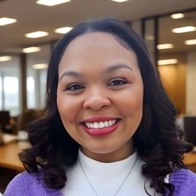 Woman with dark hair smiles, wearing a purple sweater, white top, in an office setting.