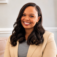 Woman with dark hair smiles, wearing a purple sweater, white top, in an office setting.