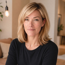 Woman with blonde hair smiling in a room; a couch and window in the background.