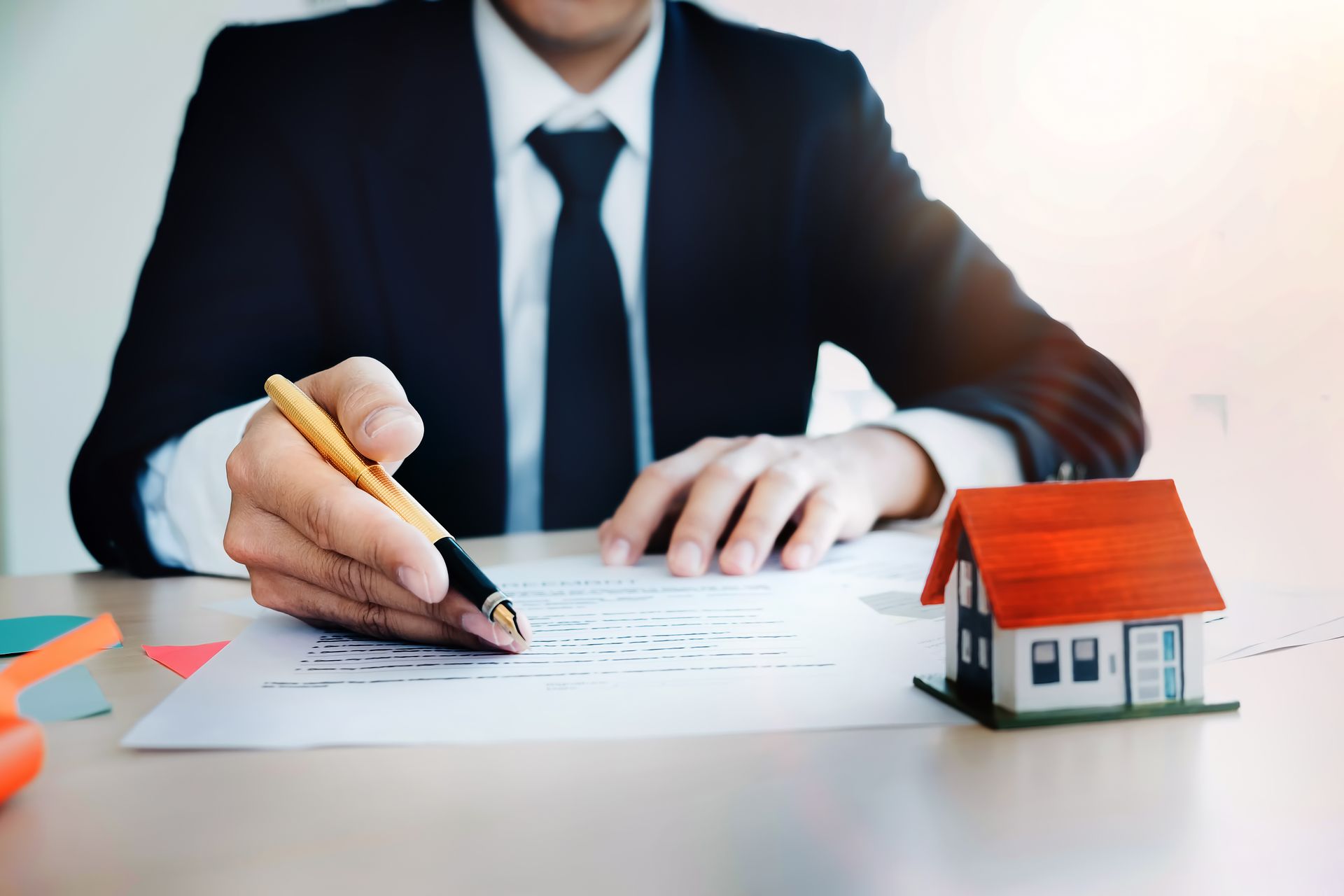 Person signing paperwork at a desk beside a small house model, suggesting a real estate. Person signing paperwork at a desk beside a small house model, suggesting a real estate.