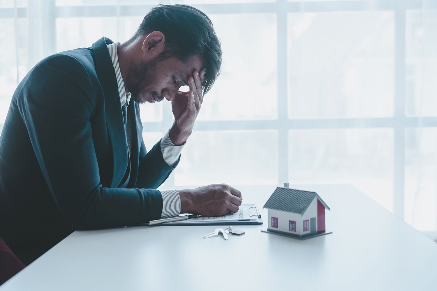 A man holding his head while seated at an office table with a model home, keys, and clipboard.