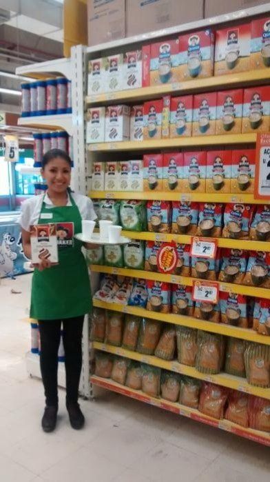 Una mujer con un delantal verde está parada frente a un estante de una tienda de comestibles sosteniendo una caja.