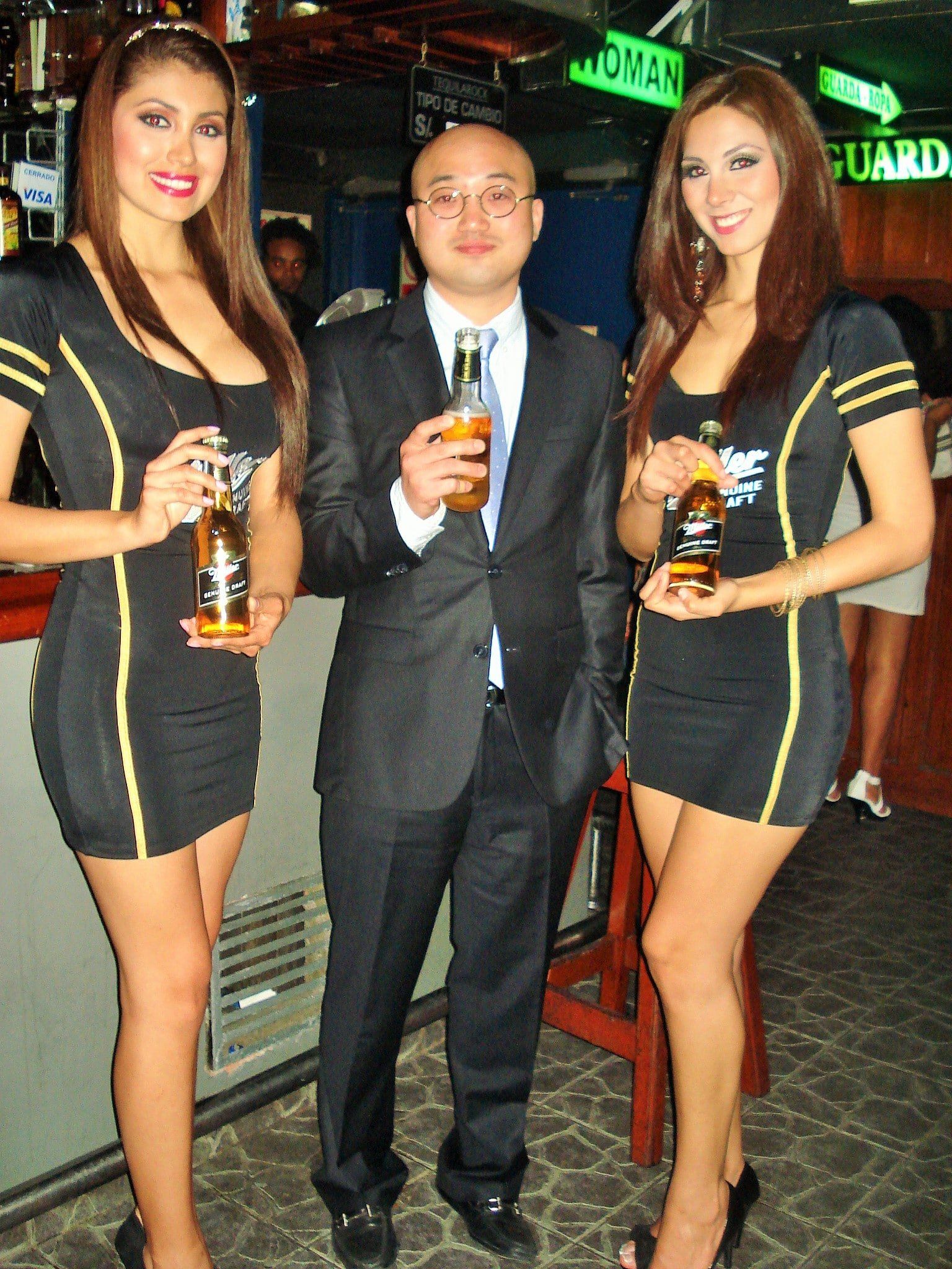 Un hombre con traje se encuentra entre dos mujeres que sostienen botellas de cerveza.