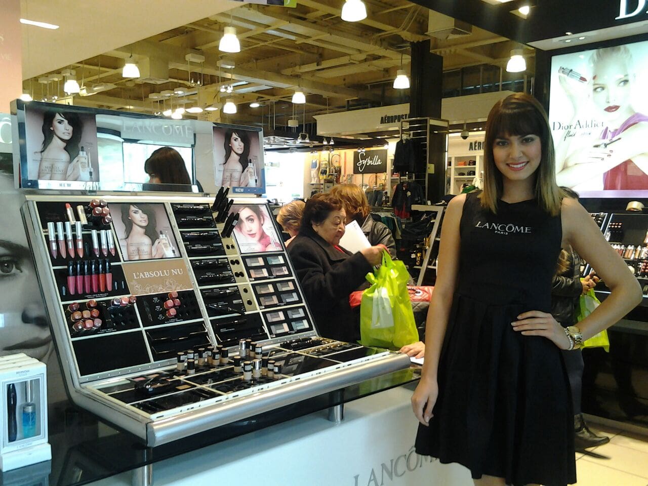 Una mujer con un vestido negro está parada frente a un expositor de maquillaje.