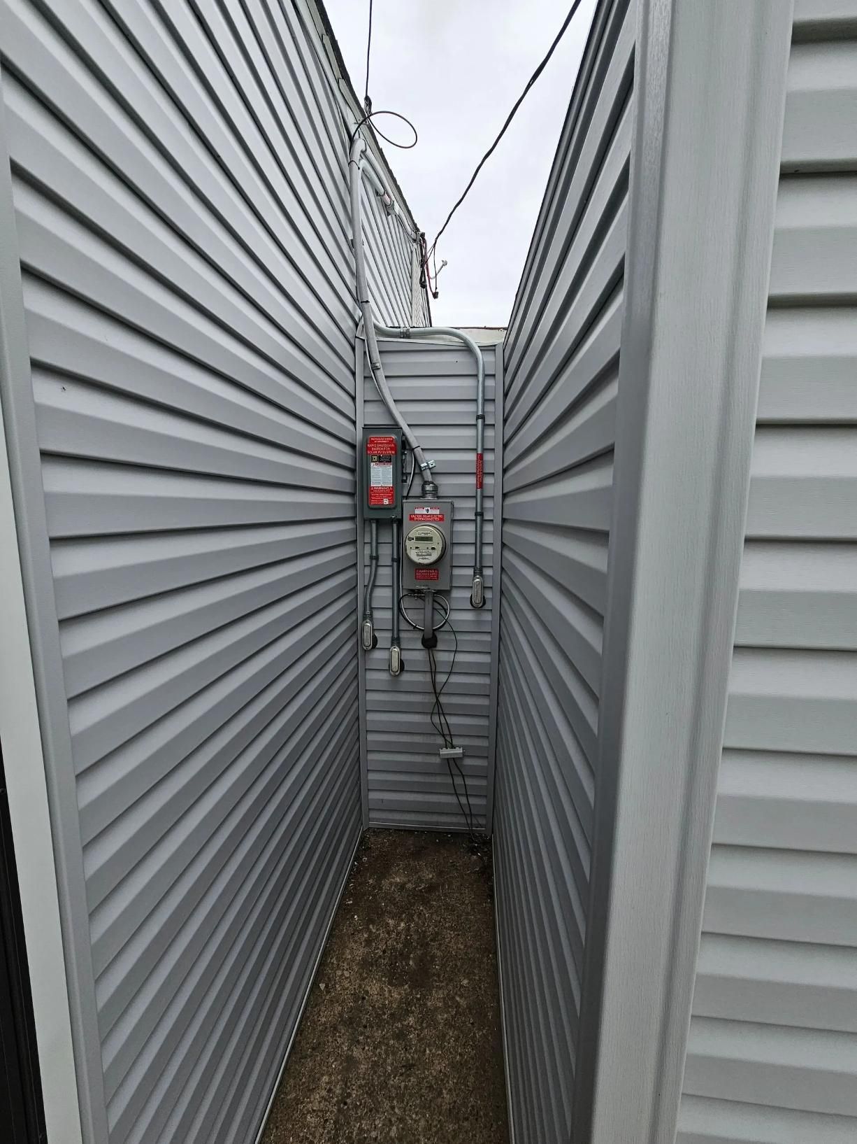 Narrow alleyway lined with gray siding, housing gas meter and electrical components.