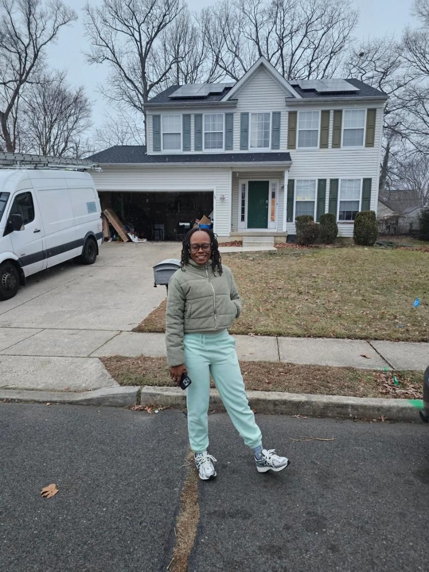 Woman in light green outfit stands in front of a white house with a garage and solar panels.