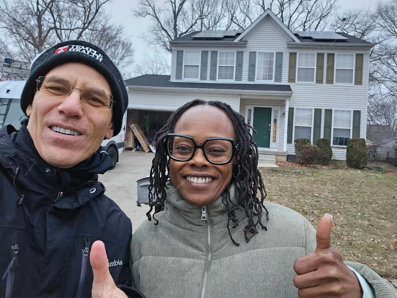 Two people giving thumbs up in front of a two-story house with solar panels on the roof.