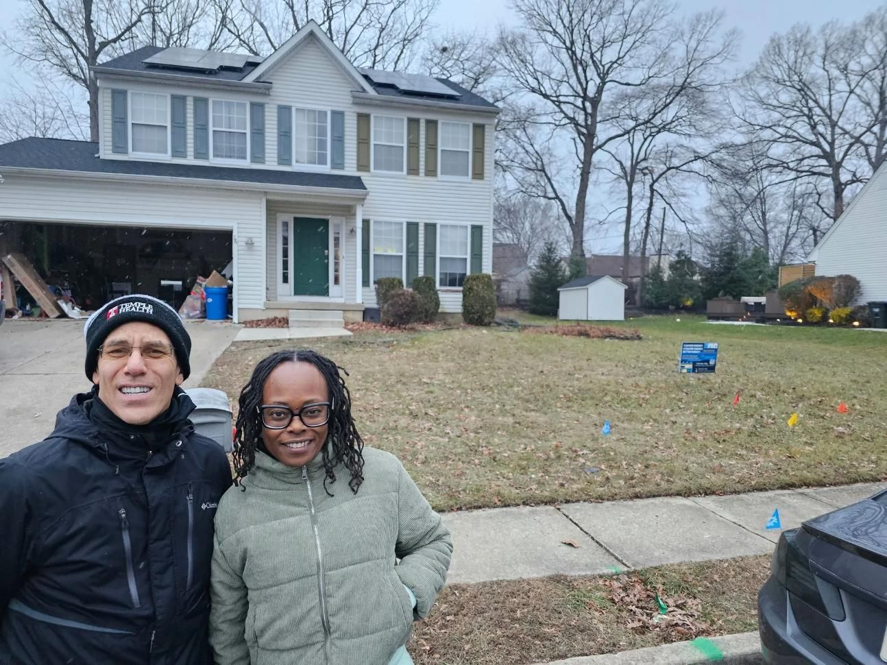 Two people standing in front of a two-story house with bare trees in the background. 