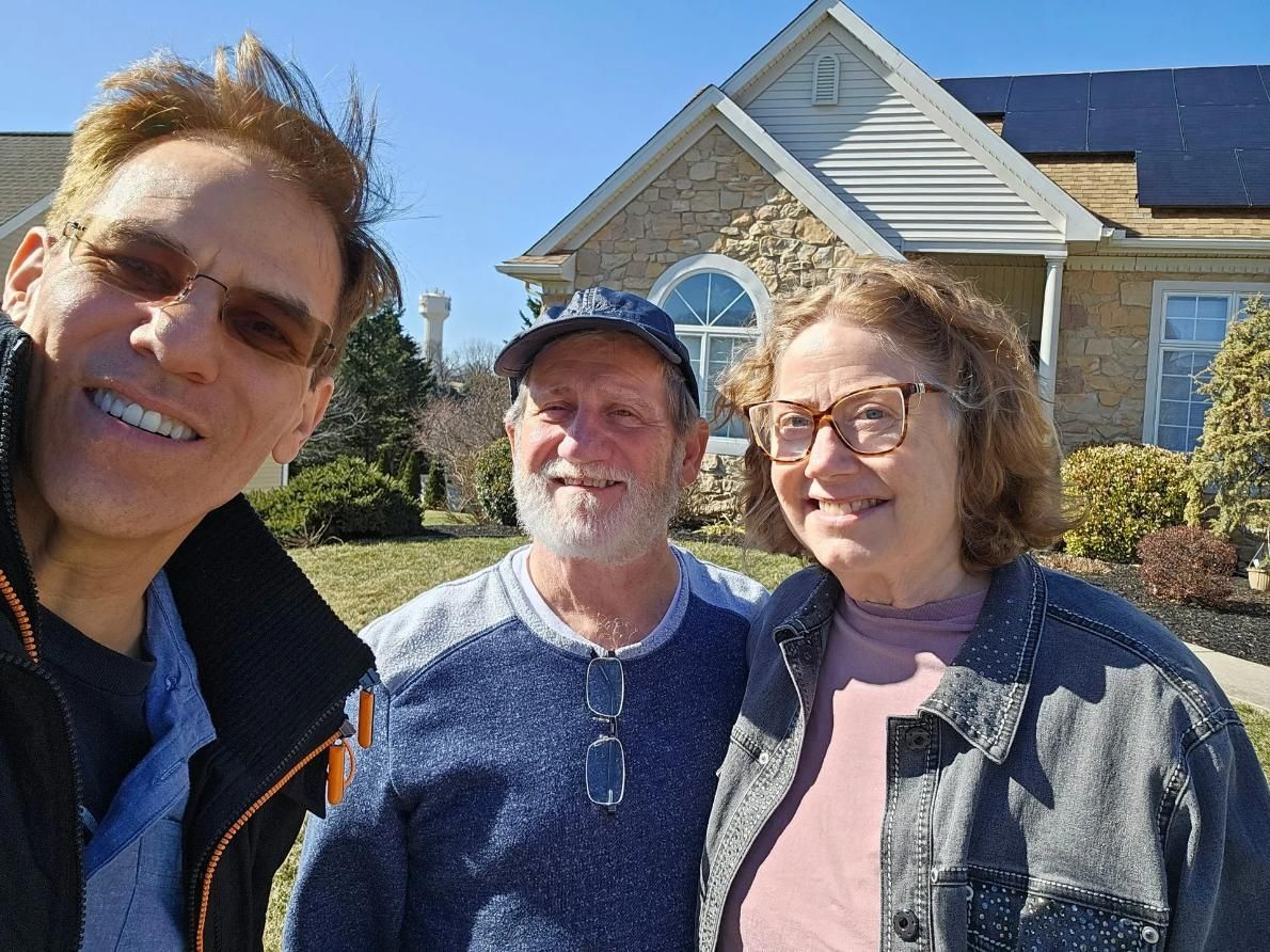 Three people smiling for a selfie outdoors in front of a house with solar panels; sunny day.