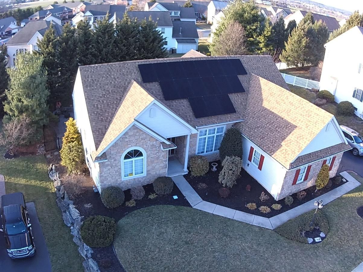 House with solar panels on roof, brown shingles, brick facade, white trim, trees, and landscaping.