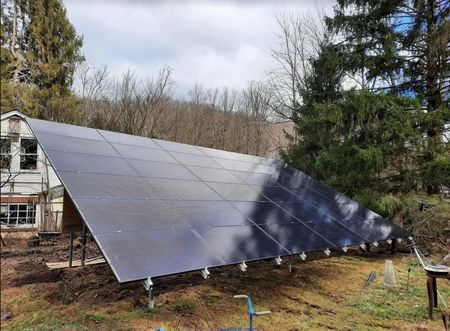 Large angled solar panel array on a grassy hillside, in front of a building and trees, cloudy sky.
