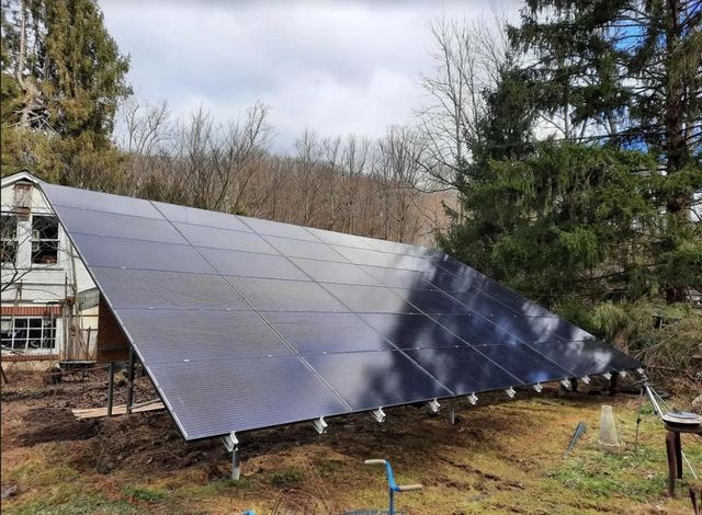 Large angled solar panel array on a grassy hillside, in front of a building and trees, cloudy sky.