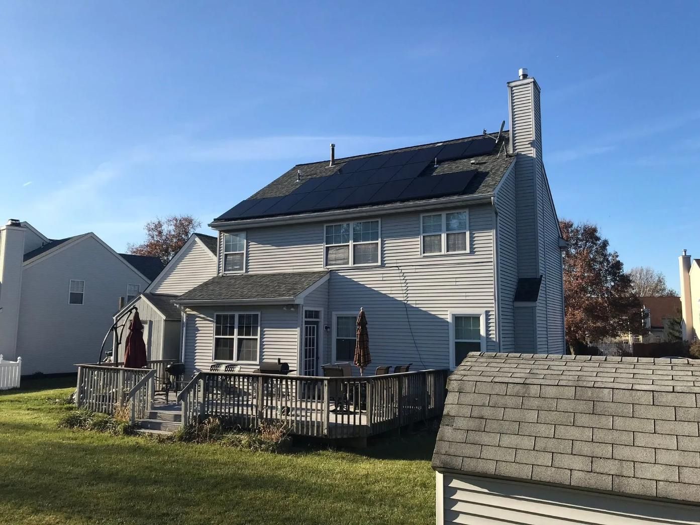 Two-story house with solar panels on roof, wooden deck, chimney, and blue sky.