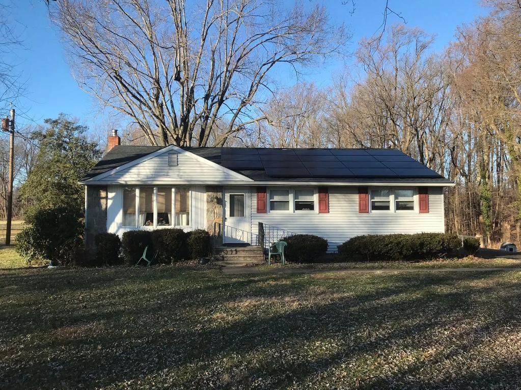Ranch-style house with solar panels on the roof. Light siding, red shutters, and a lawn with brown grass.