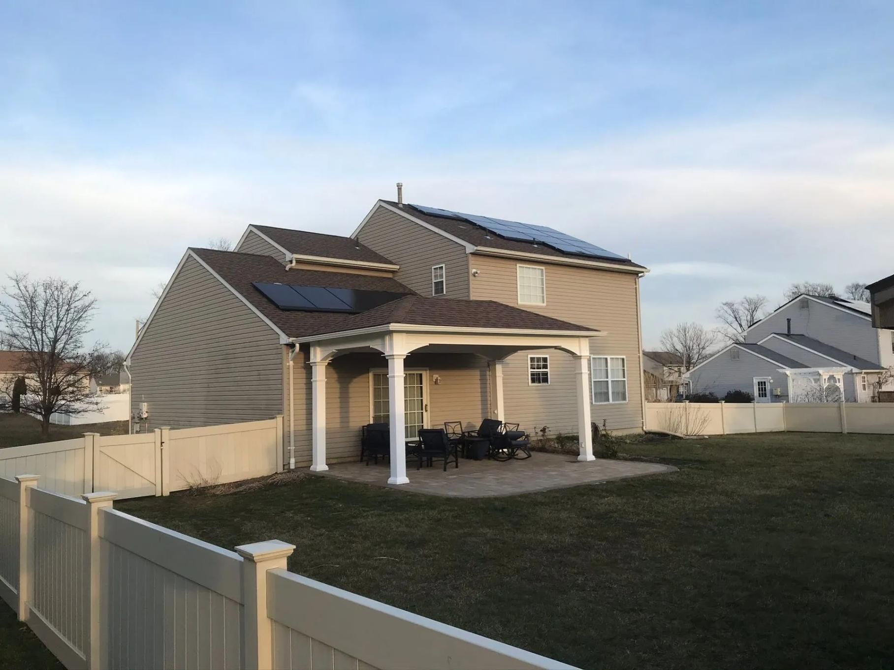 Backyard view of a two-story house with solar panels on the roof and a patio with a white fence.