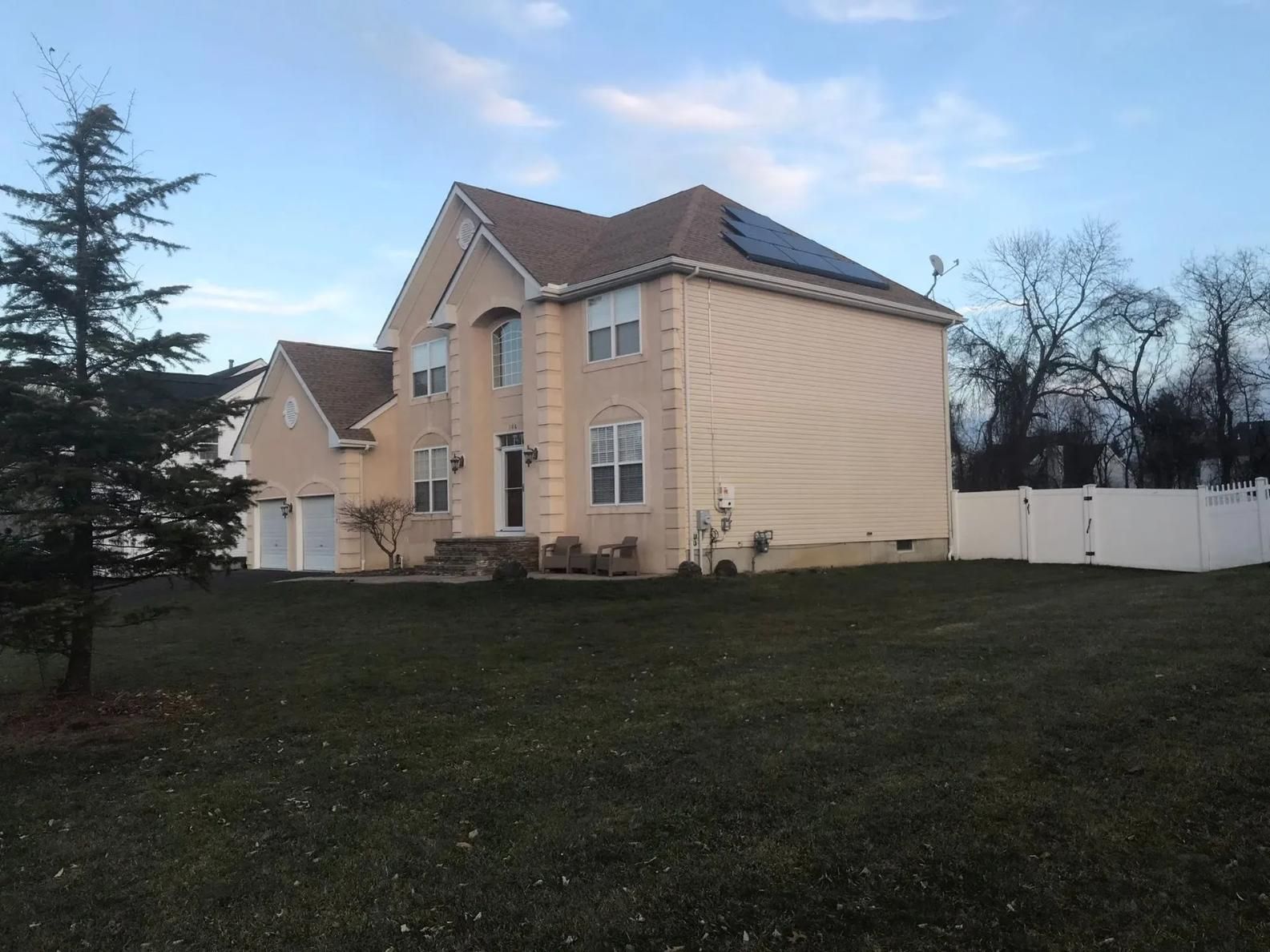 Two-story beige house with solar panels on roof; set on a lawn with a white fence and bare trees in the background.